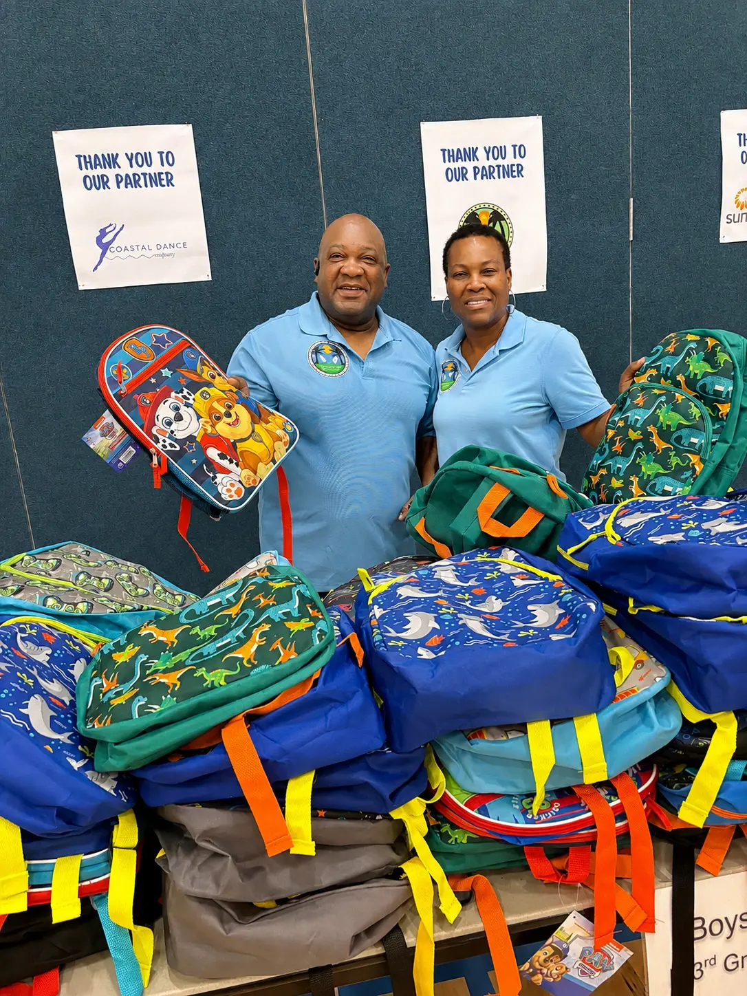 Two volunteers holding backpacks behind a table of donated bags