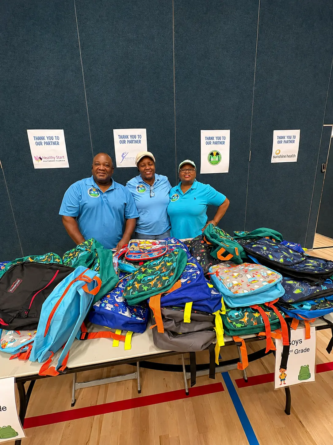 Three volunteers standing behind a table piled with backpacks