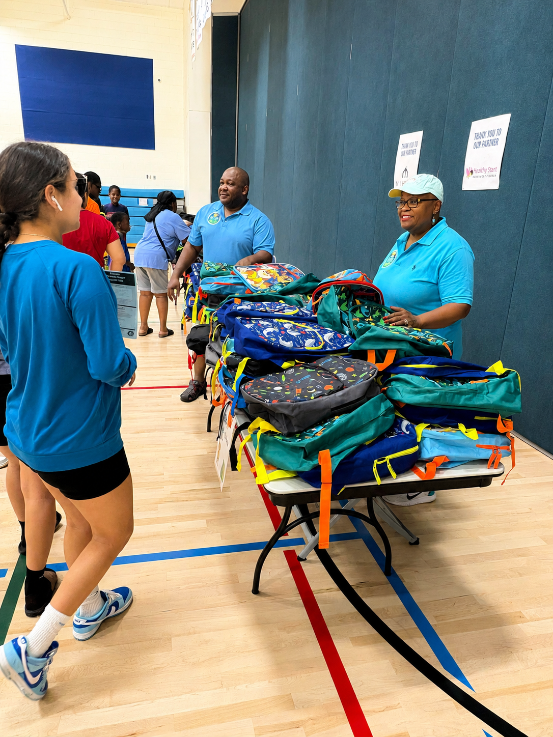 Volunteers standing behind tables of backpacks in the gym